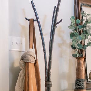 May include: A black metal coat rack shaped like tree branches with a brown leather tote bag hanging from one of the branches. The rack is standing on a white surface next to a vase with green leaves.