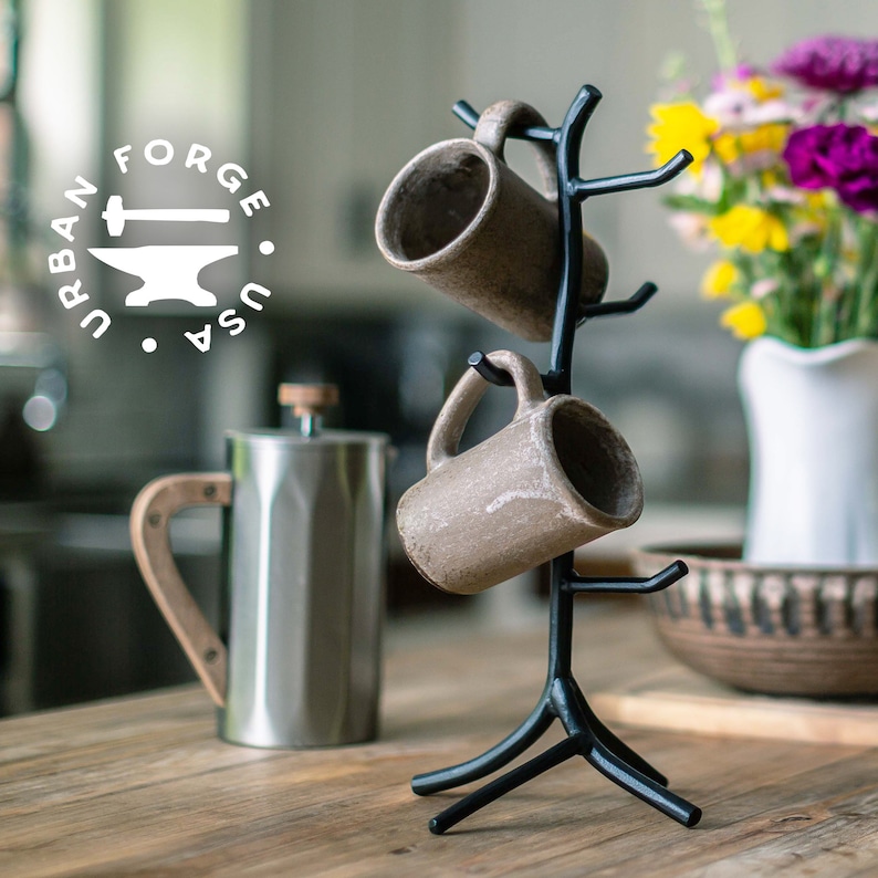 A black hand forged iron mug tree with two brown ceramic mugs hanging from it. The mug tree has a tree branch design with a wide base. The mugs are hanging from the branches of the tree. The mug tree is on a wooden table.