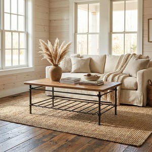 May include: A living room scene featuring a light-colored sofa with beige pillows and a woven throw. A rectangular wooden coffee table with a black metal frame sits on a jute rug. A vase with dried pampas grass is on the table.