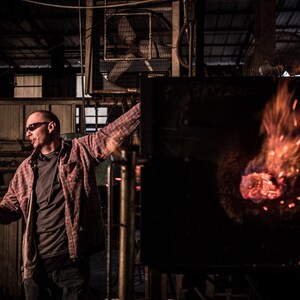 May include: A man wearing a red and black plaid shirt and sunglasses stands in a dark industrial setting. He is looking to the left, and there is a large furnace with flames and glowing embers in the background.