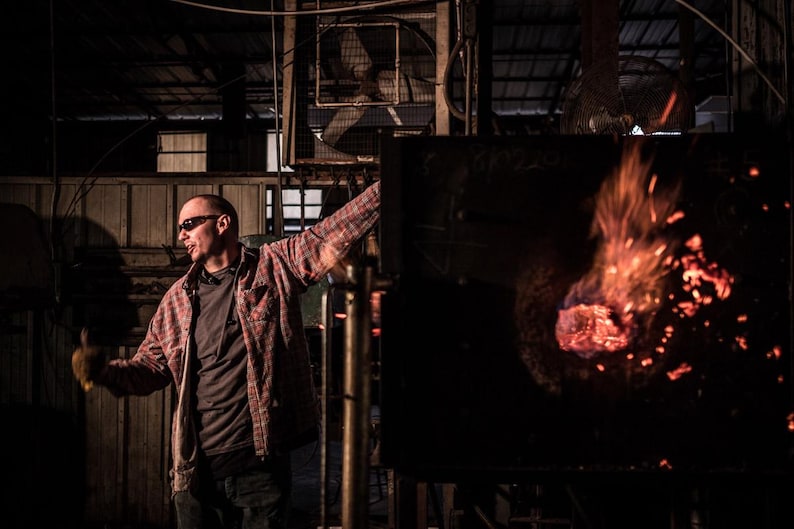 May include: A man wearing a red and black plaid shirt and sunglasses stands in a dark industrial setting. He is looking to the left, and there is a large furnace with flames and glowing embers in the background.
