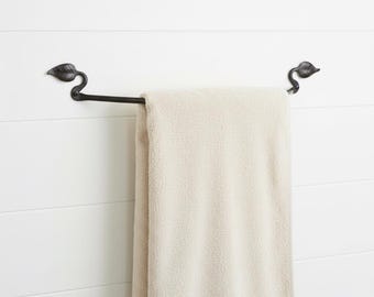 May include: A black metal towel bar with leaf-shaped accents on each end, holding a beige towel. The bar is mounted on a white, horizontal-slatted wall, creating a simple, elegant bathroom accessory.
