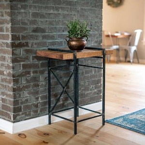 May include: A rustic side table with a wooden top and a black metal frame. The table features a small copper pot with greenery on top. The table is placed next to a gray brick wall and a light wood floor.