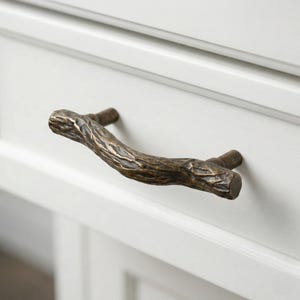 May include: A bronze-colored cabinet pull in the shape of a tree branch. The textured surface of the pull is detailed with ridges and knots, giving it a natural, rustic appearance. The pull is mounted on a white cabinet door.