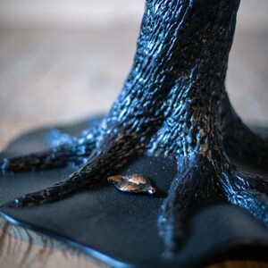 A black tree metal table base with a textured surface. The base of the sculpture is a flat, black surface with a small, gold leaf on it.