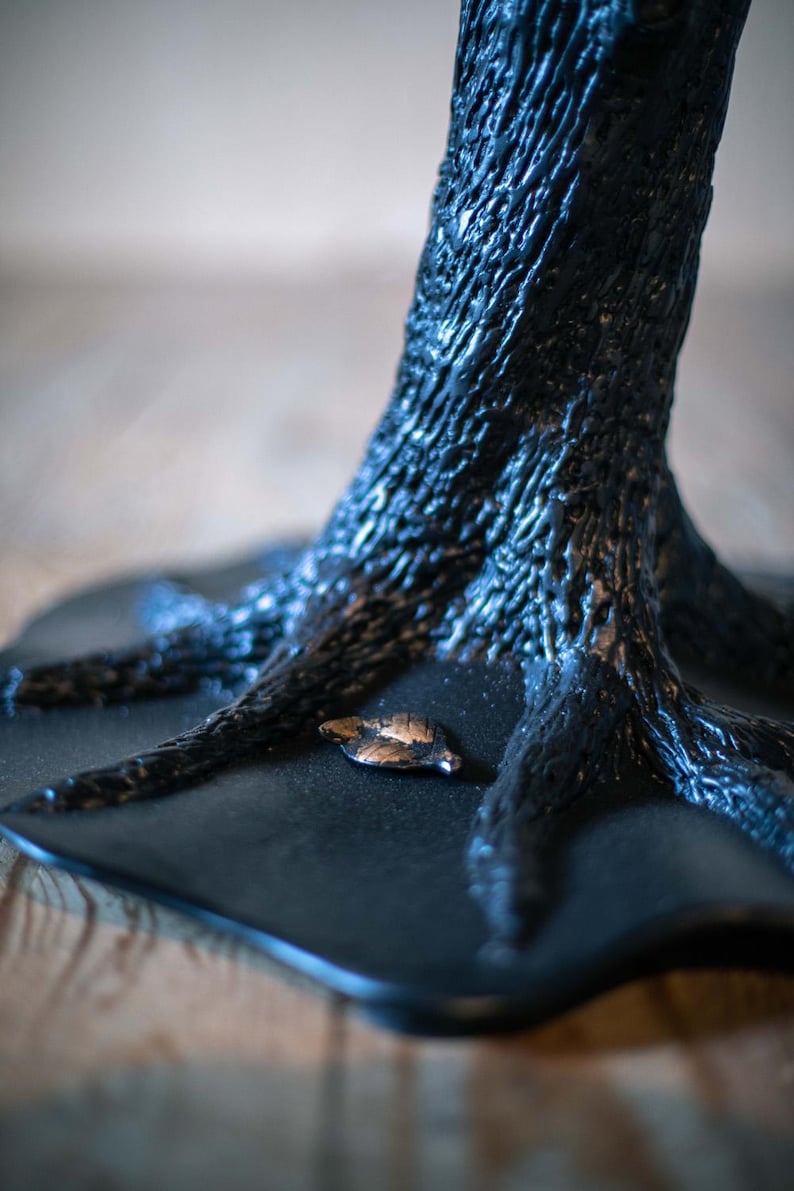 A black tree metal table base with a textured surface. The base of the sculpture is a flat, black surface with a small, gold leaf on it.