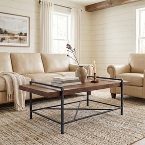May include: A modern living room scene featuring a brown wooden coffee table with a black metal frame. The table holds books and a vase with dried plants. A beige leather sofa and armchair are in the background, with a neutral rug.