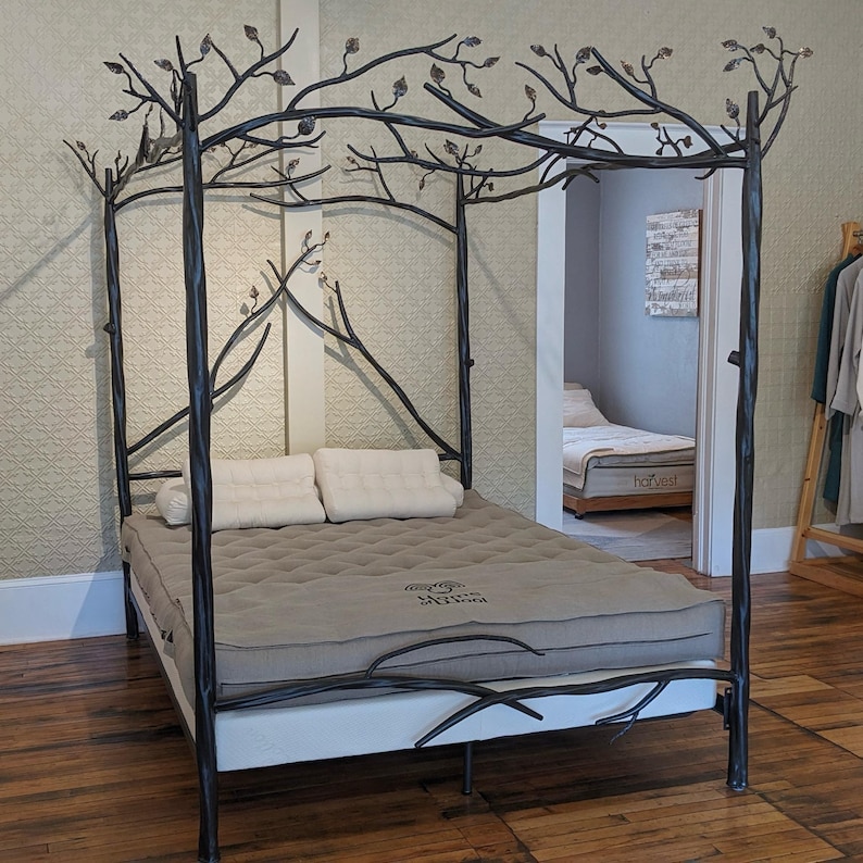 A black hand forged iron canopy bed frame with a tree branch design. The bed has a gray mattress with a quilted pattern and a white pillow. The bed is in a room with wood floors and a light-colored wall.
