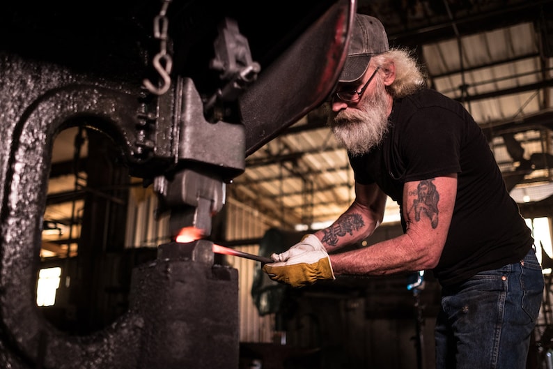 A blacksmith using a large black metal press to shape a piece of hot metal. The metal is glowing red hot and is being pressed into a flat shape.