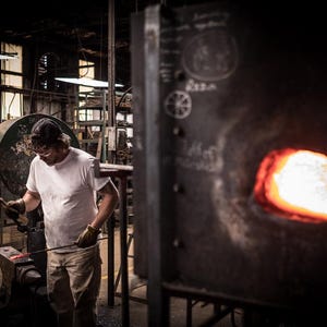 May include: A person wearing a white t-shirt and a baseball cap works with a metal rod in a workshop. The rod is being heated in a forge with a bright orange glow.