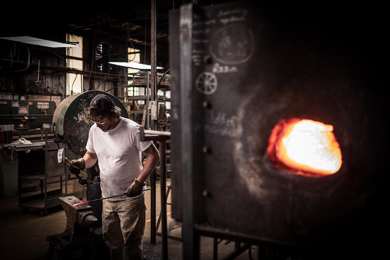 May include: A person wearing a white t-shirt and a baseball cap works with a metal rod in a workshop. The rod is being heated in a forge with a bright orange glow.