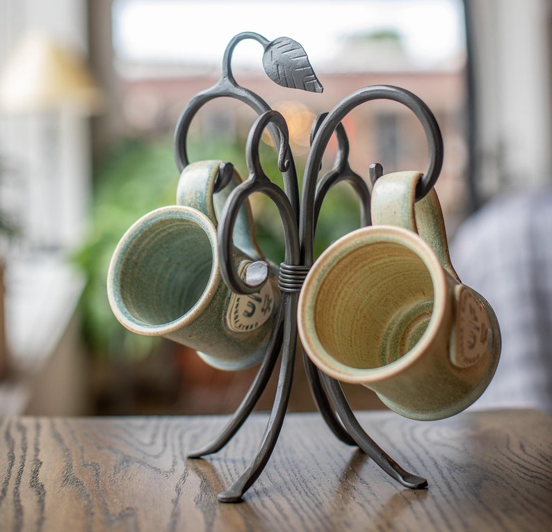 A black metal mug rack with a leaf design. The rack is holding two ceramic mugs with green and brown glaze.
