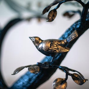 A metal bird sculpture with gold accents perched on a black metal tree branch with leaves.