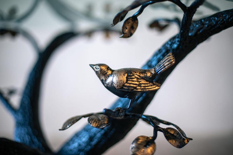 A metal bird sculpture with gold accents perched on a black metal tree branch with leaves.