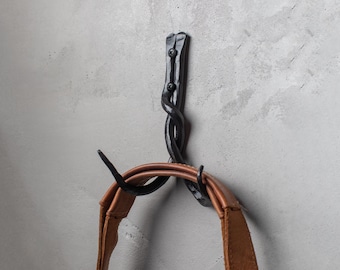 May include: A black, wrought iron wall hook with a twisted design. The hook is mounted on a gray wall and holds a brown leather strap. The hook is secured with two black screws.