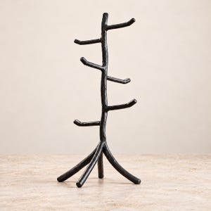 May include: A black metal jewelry stand shaped like a tree with a central trunk and multiple branches extending outward. The stand features a textured, uneven surface and is supported by three sturdy legs at the base for stability.