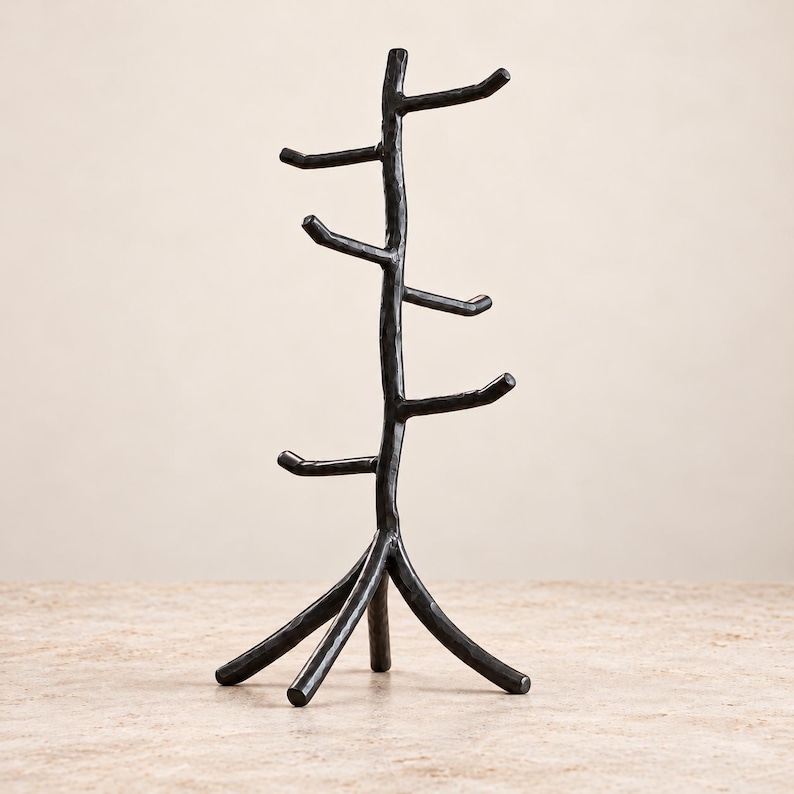 May include: A black metal jewelry stand shaped like a tree with a central trunk and multiple branches extending outward. The stand features a textured, uneven surface and is supported by three sturdy legs at the base for stability.