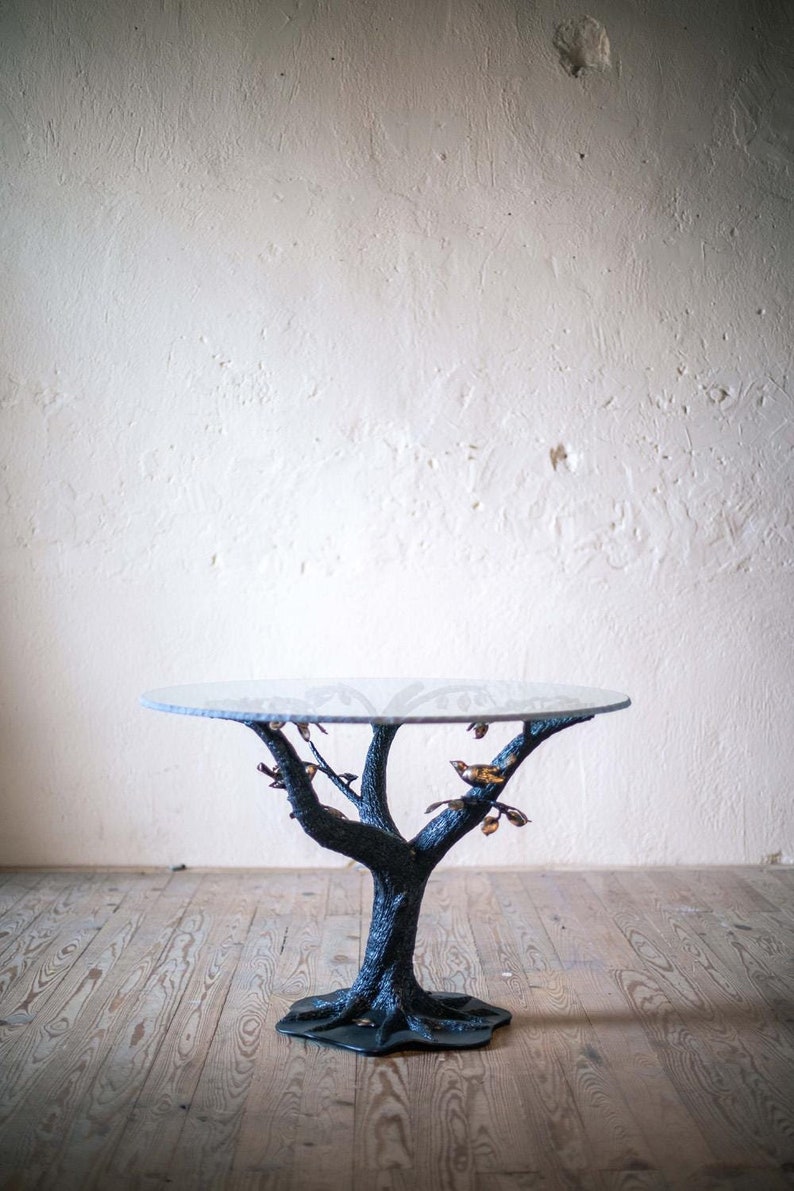 A round glass table top sits on a black metal tree trunk base with gold leaf branches and birds. The table is on a wooden floor.