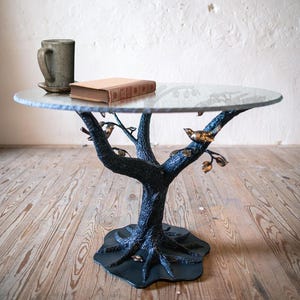 A round glass table top with a black metal tree-shaped base. The base has a textured bark-like finish and gold-colored birds perched on the branches. The table is on a wooden floor.