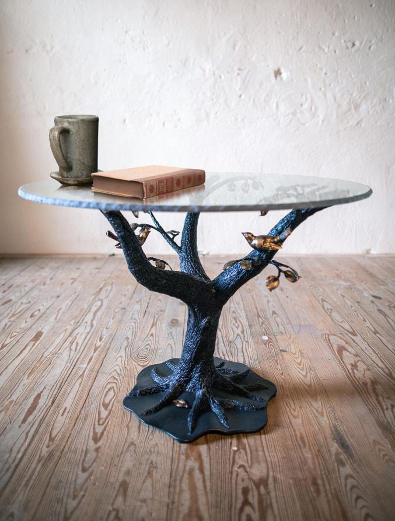 A round glass table top with a black metal tree-shaped base. The base has a textured bark-like finish and gold-colored birds perched on the branches. The table is on a wooden floor.