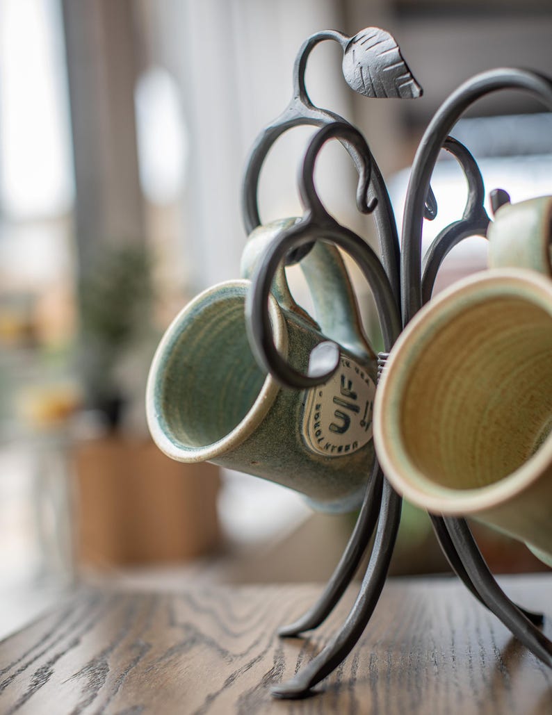 A metal mug holder shaped like a tree branch with leaves. The holder has four hooks and is holding two green ceramic mugs.