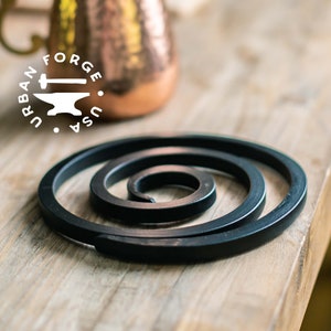 May include: A black metal spiral trivet, also known as a pot holder, is on a wooden surface. The trivet is made of a single piece of metal that is coiled into a spiral shape. The trivet is designed to protect surfaces from heat.
