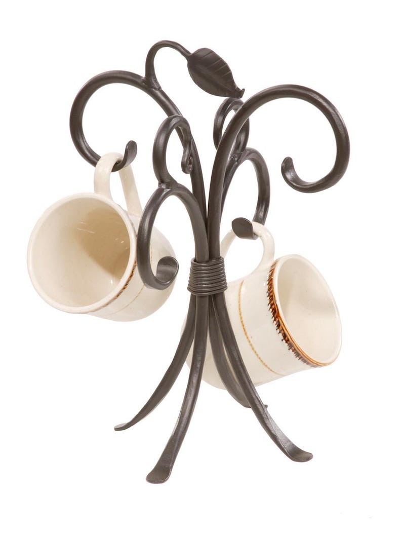 A black metal mug tree with four hooks. The mug tree has a decorative leaf design and a base with four legs. Two white mugs with brown designs are hanging on the hooks.
