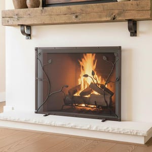 May include: A decorative fireplace screen with a black metal frame and a mesh center. The screen features a stylized vine design with leaves. A fire is burning brightly behind the screen, with flames and logs visible.