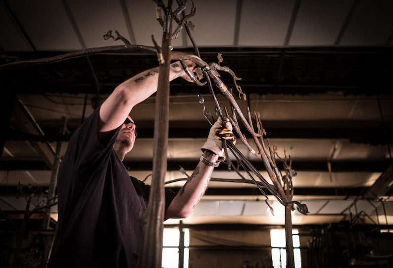 K&ouml;nnte beinhalten: Eine dunkelbraune Metallskulptur in Form eines Baumes mit d&uuml;nnen, strukturierten &Auml;sten. Die Metallstruktur weist eine raue, organische Oberfl&auml;che mit mehreren nach oben ragenden Gliedern auf, gefertigt aus dunklem, verwittertem Eisenmaterial.