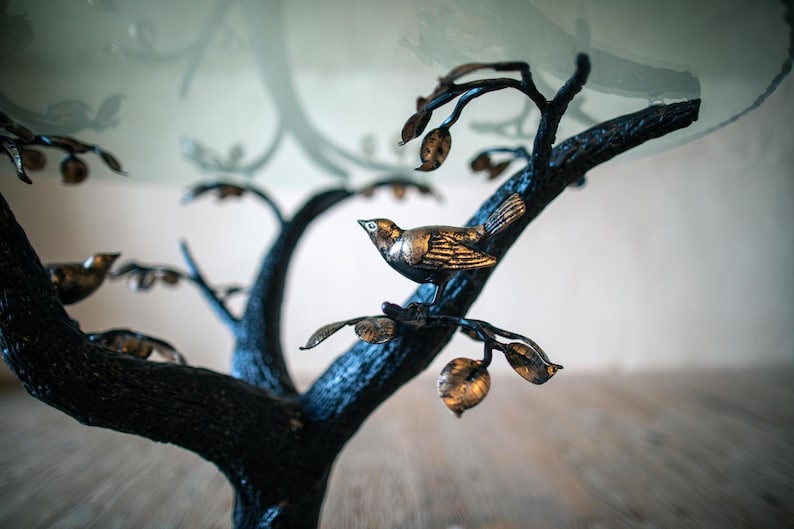A black metal tree table with a glass top. The tree has branches with small gold leaves and birds perched on them.