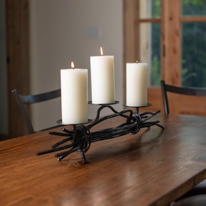 May include: A black metal candelabra with three lit white pillar candles. The candelabra has a rustic, twisted design and sits on a wooden table. The candles are approximately 4 inches in diameter.