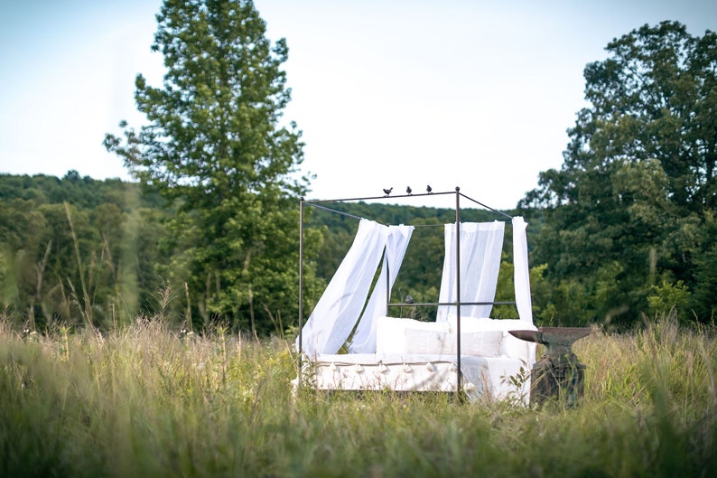 A black canopy bed frame with white sheer curtains in a grassy field. The bed frame is made of metal and has a simple design. The bed is made up with white linens and pillows. The bed is surrounded by tall green grass and trees in the background.
