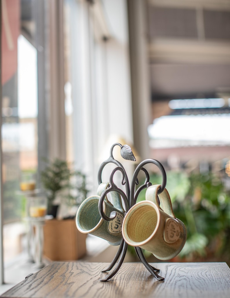 Two ceramic mugs with green and beige glaze hanging on a black metal mug tree.