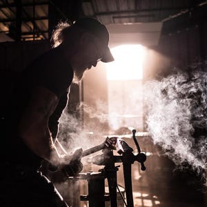 May include: A person wearing a black cap and a black shirt is working with a metal object in a workshop. The object is surrounded by smoke and steam.