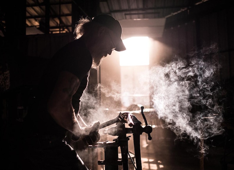 May include: A person wearing a black cap and a black shirt is working with a metal object in a workshop. The object is surrounded by smoke and steam.