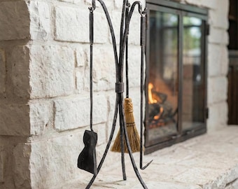 May include: A black wrought iron fireplace tool set with a stand. The set includes a poker, shovel, and broom. The tools are displayed on a stone hearth in front of a fireplace.