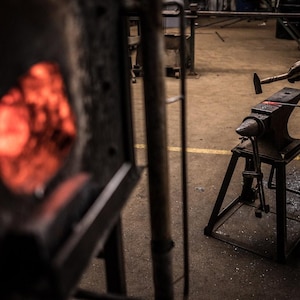 May include: A blacksmith uses a hammer to strike a piece of metal on an anvil. The metal glows red hot in a nearby forge.