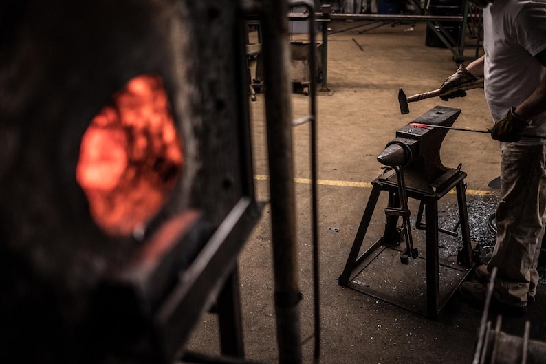 May include: A blacksmith uses a hammer to strike a piece of metal on an anvil. The metal glows red hot in a nearby forge.