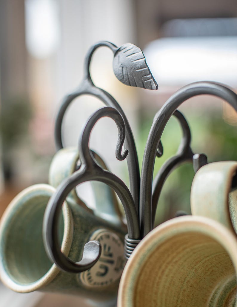 A black metal mug rack with a leaf design. The rack is holding several green and cream colored mugs.