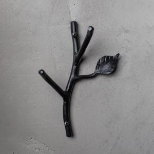 May include: A black metal wall hook in the shape of a tree branch with a leaf. The hook has three prongs for hanging items. The branch is mounted on a gray wall.