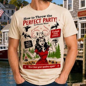 May include: A cream-colored short-sleeve t-shirt featuring a graphic print with the text "How to Throw the PERFECT PARTY! by Patricia Moyer". The illustration depicts a person in a red dress, a fox, birds, trees, and text boxes reading "Give the PERFECT TOAST!" and "Enchant YOUR GUESTS!" with "Never have your qualms again!" at the bottom.