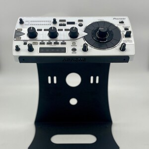 Op de afbeelding: Een witte Pioneer DJ-mixer met een zwarte standaard. De mixer heeft een grote centrale knop en verschillende kleinere knoppen en knoppen. De standaard is gelabeld "APACHE".