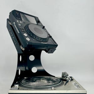 Pode incluir: Um leitor multimédia DJ Pioneer preto montado num suporte de metal preto sobre um gira-discos prateado. O suporte apresenta recortes circulares e a palavra APACHE impressa na lateral. O leitor possui uma roda central e vários botões.