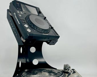 May include: A black Pioneer DJ media player mounted on a black metal stand above a silver turntable. The stand features circular cutouts and the word APACHE printed on the side. The media player has a central jog wheel and various buttons.