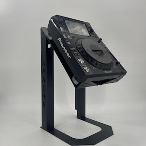 May include: A black metal DJ equipment stand with a vertical support arm featuring the laser-cut text APACHE. The stand holds a black Pioneer digital media player at an angled position, showcasing a central jog wheel, display screen, and buttons.