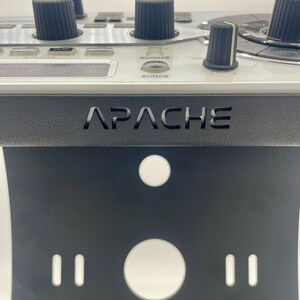 Op de afbeelding: Een zwarte standaard met het woord "APACHE" erop gedrukt. De standaard heeft twee kleine gaten en een groot gat aan de voorkant. De standaard is ontworpen om een ​​muziekproductieapparaat te houden.