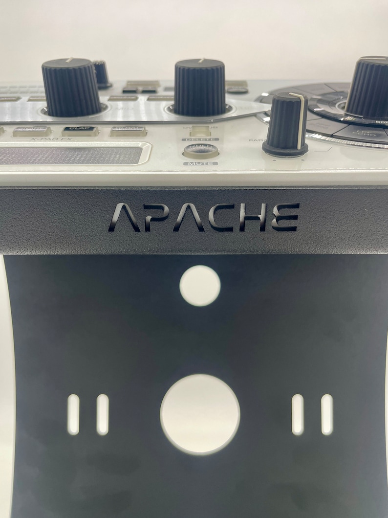 Op de afbeelding: Een zwarte standaard met het woord "APACHE" erop gedrukt. De standaard heeft twee kleine gaten en een groot gat aan de voorkant. De standaard is ontworpen om een ​​muziekproductieapparaat te houden.