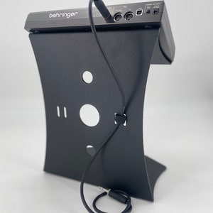 Op de afbeelding: Zwarte Behringer audio-interface met een zwarte standaard en een zwarte audiokabel die aan de achterkant is aangesloten.
