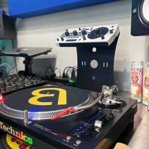 Op de afbeelding: Een zwarte draaitafel met een zilveren toonarm en een zwarte mat met een gele letter 'a' erop. De draaitafel staat op een zwart oppervlak. Achter de draaitafel staat een witte DJ-mixer op een zwarte standaard.