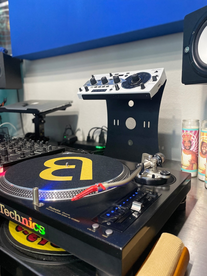 Op de afbeelding: Een zwarte draaitafel met een zilveren toonarm en een zwarte mat met een gele letter 'a' erop. De draaitafel staat op een zwart oppervlak. Achter de draaitafel staat een witte DJ-mixer op een zwarte standaard.